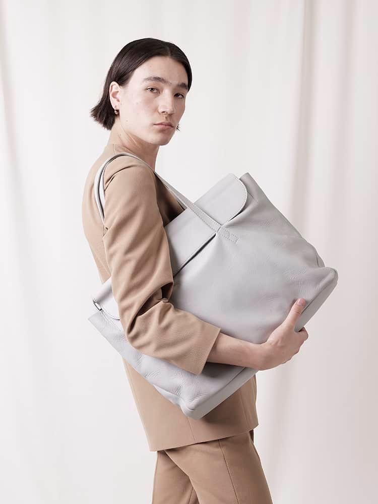 Person in beige suit holding large light gray leather tote bag against soft background
