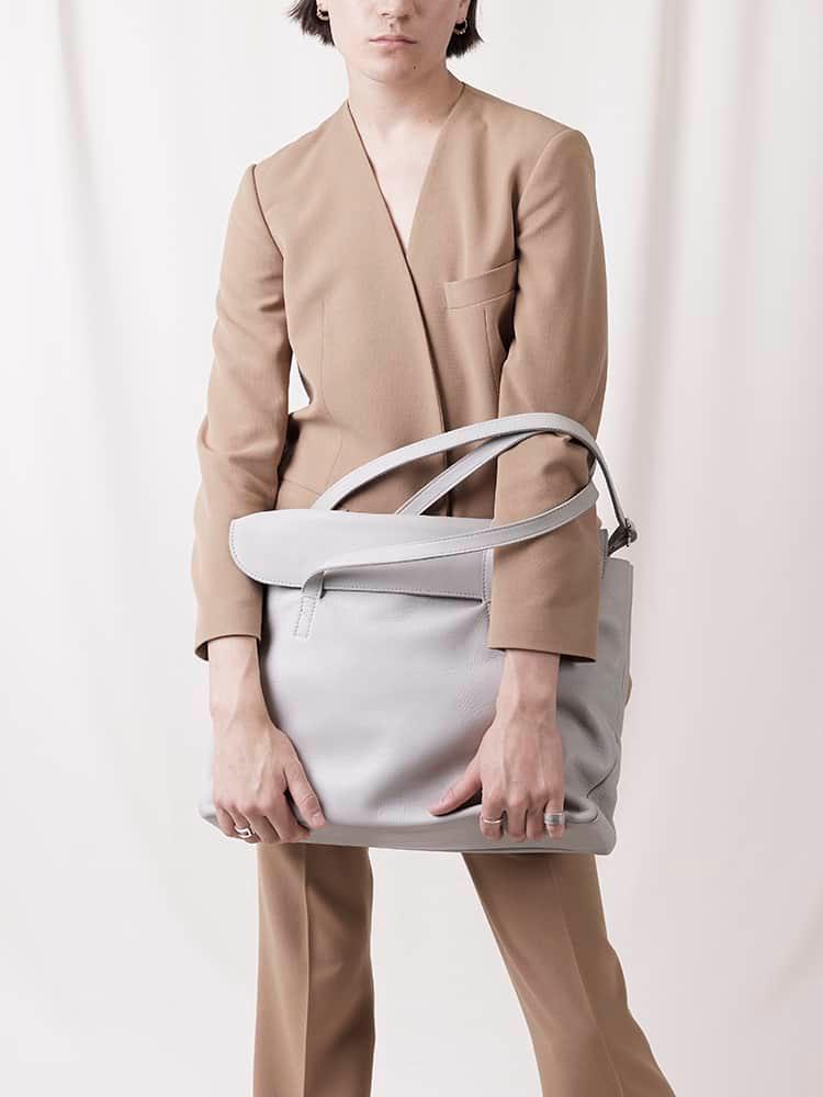 Woman in tan suit holding large light grey leather shoulder bag against neutral background