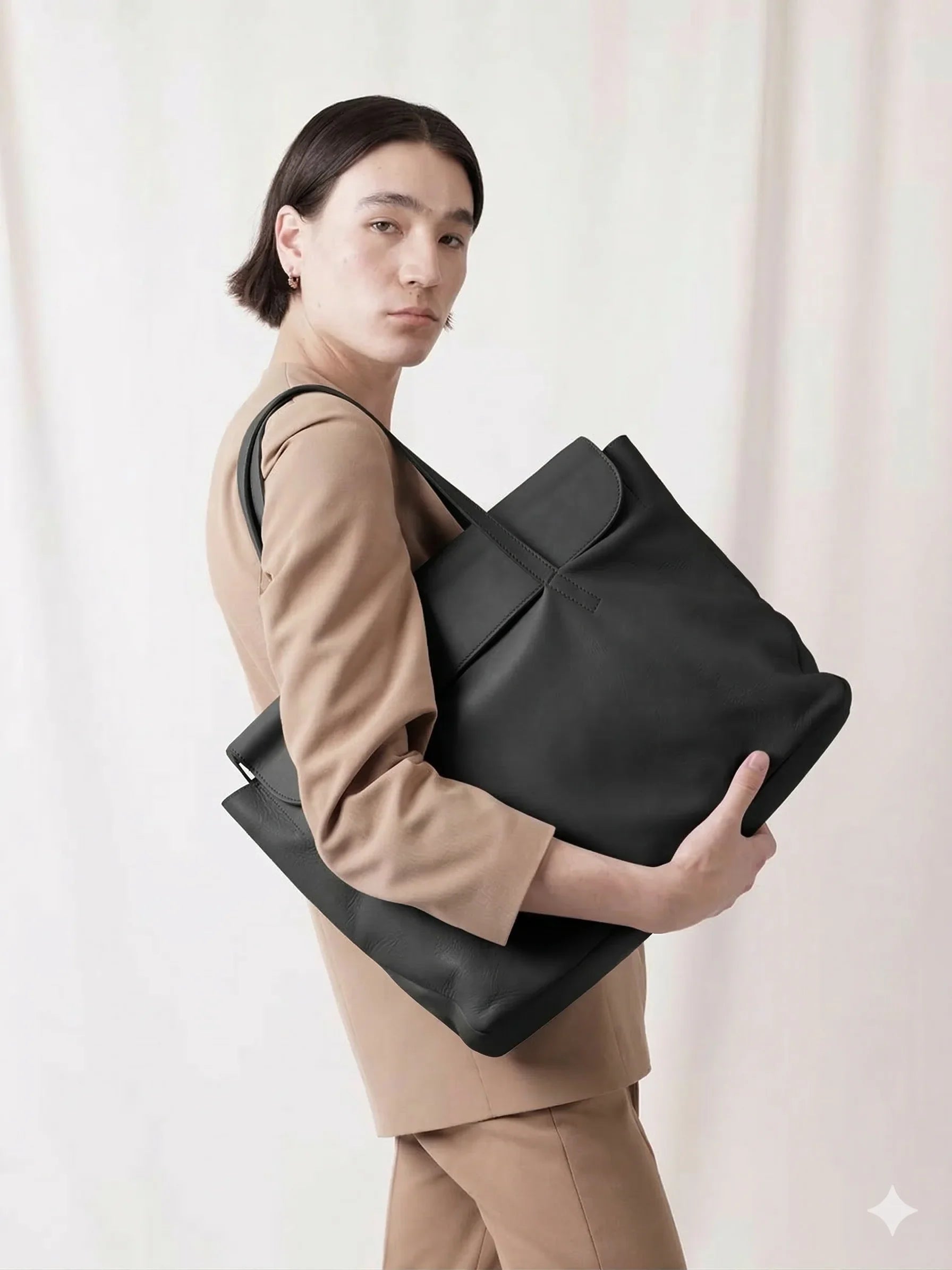 Person in beige suit carrying large black leather tote bag against neutral background