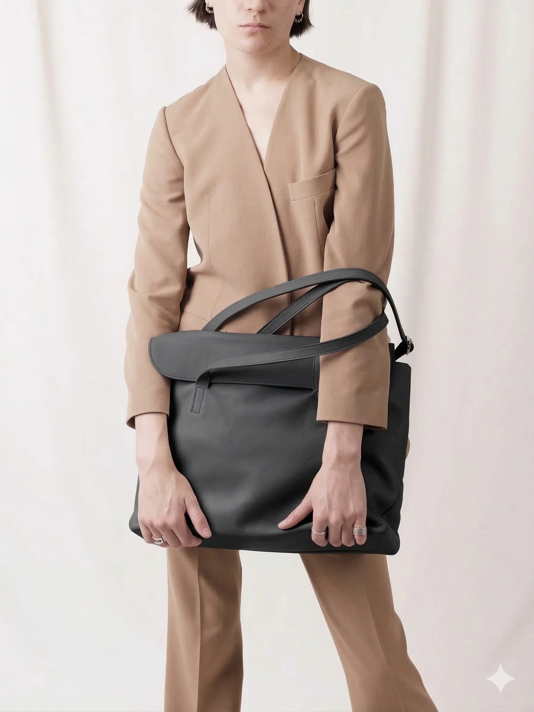 Woman in tan suit holding large black leather tote bag against neutral background
