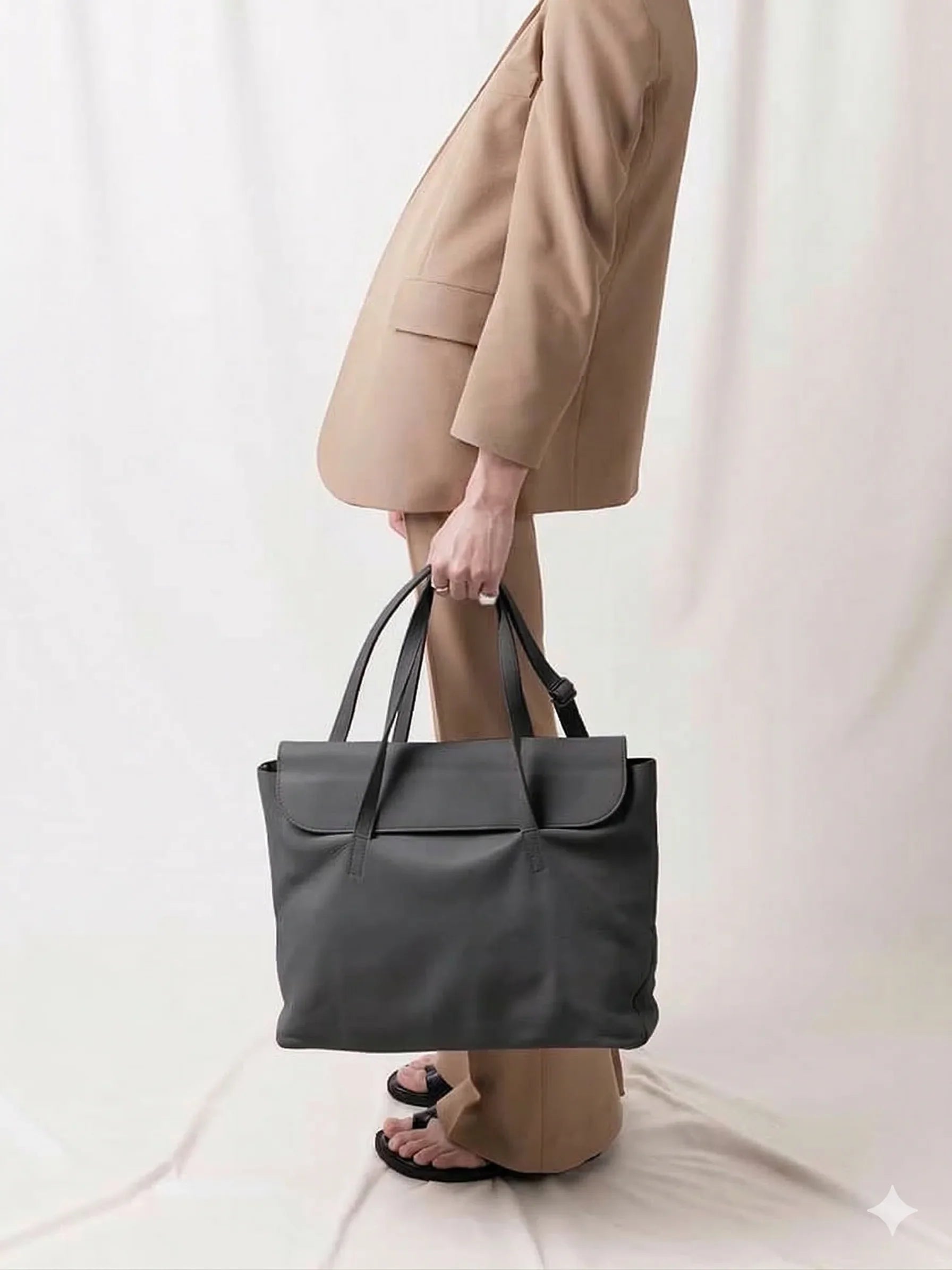 Person in beige suit holding a large black leather tote bag against a neutral background