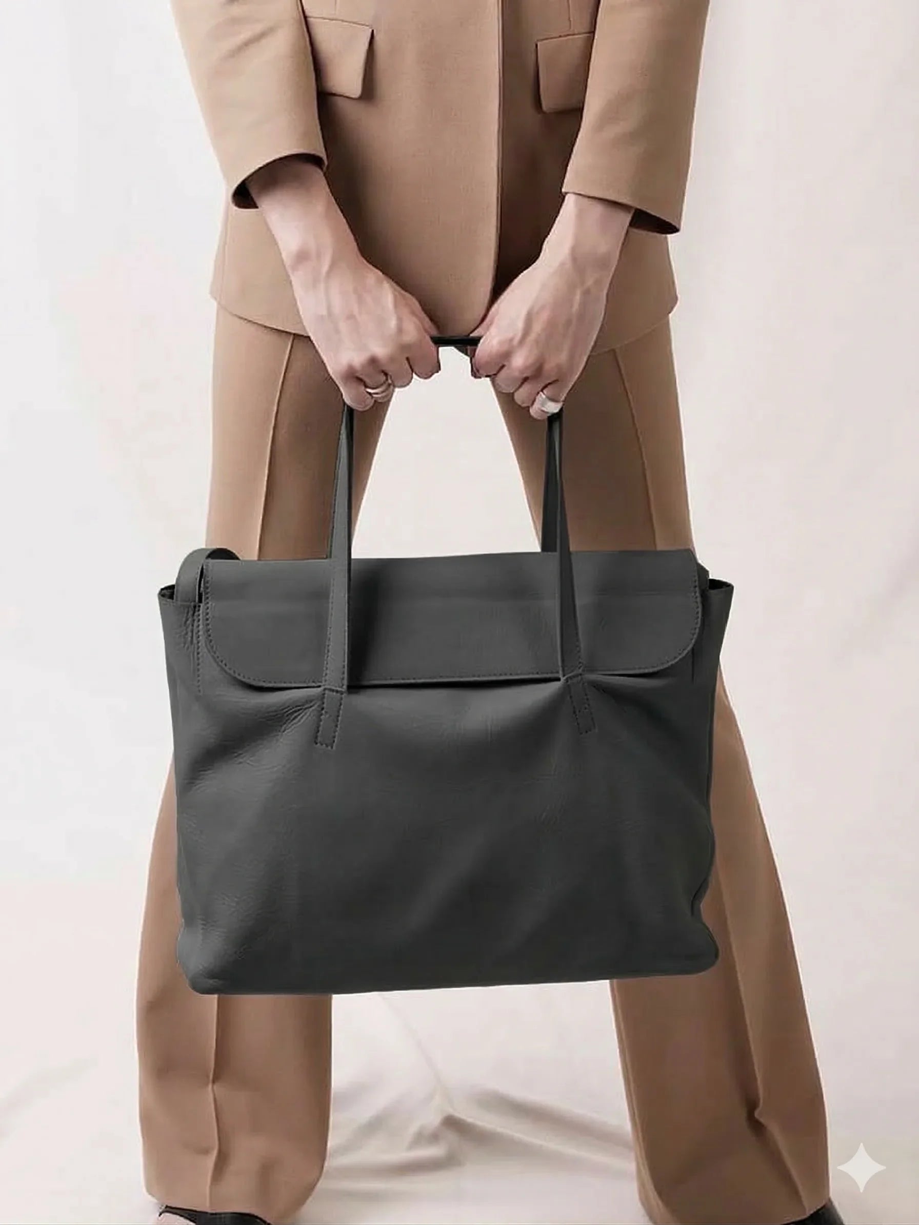 Handcrafted black leather tote bag held by a person in a tan suit, minimalist background